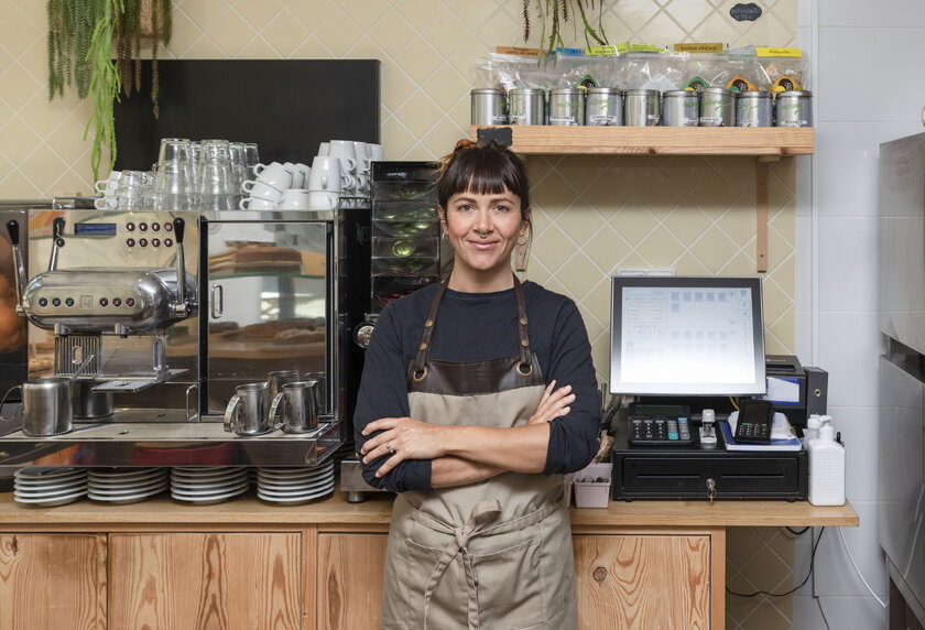 Junge Unternehmerin mit verschraenkten Armen hinter dem Tresen ihres Coffee Shops.