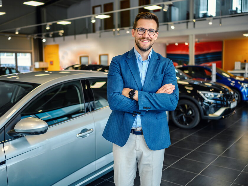 Mann in legerem Business Outfit steht selbstbewusst in Ausstellungsraum vor einer Vielzahl an Autos.