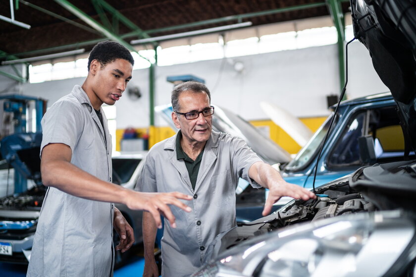 Automechaniker-Meister bespricht mit jungem Mitarbeiter Details des Automotors in der Werkstatt.